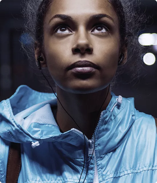 Portrait en gros plan d'une jeune athlète portant un coupe-vent bleu et des écouteurs, le regard levé vers le haut en pleine concentration nocturne.